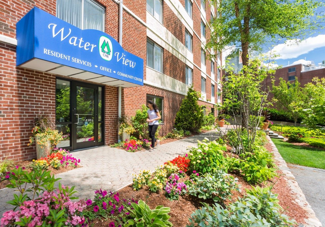 Person walking along a brick apartment entrance with a blue Water View sign and flowering landscaping.