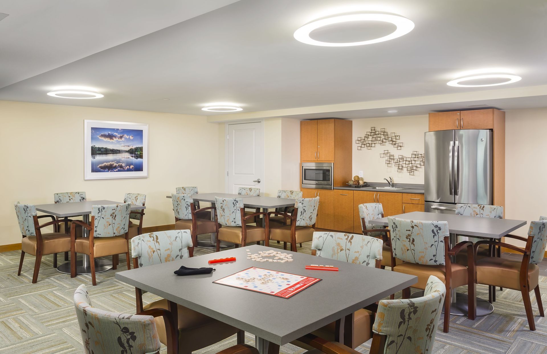 A brightly lit, neutral-toned communal room featuring several square tables with patterned chairs, a fridge, and art.