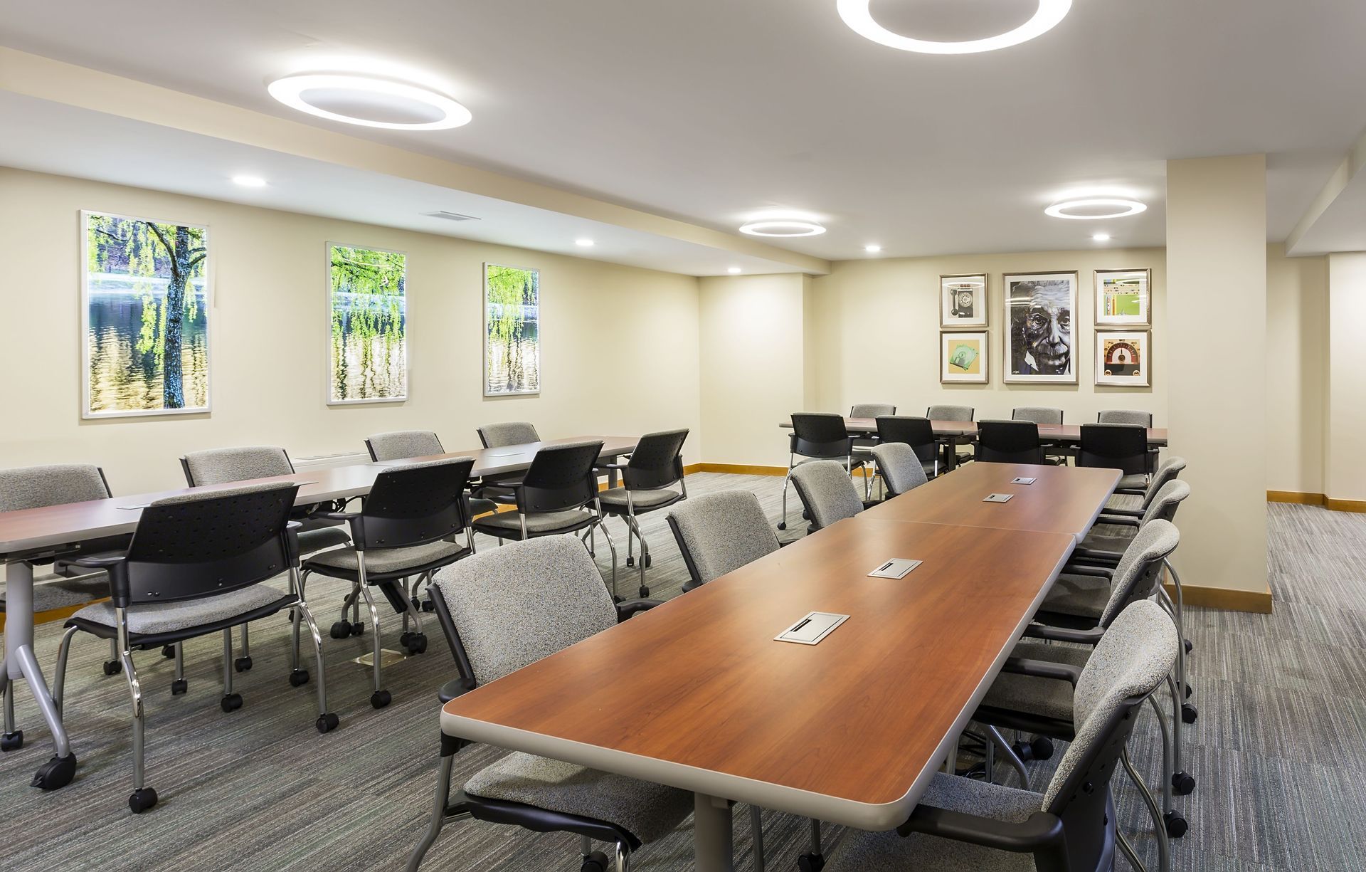 A meeting room with long wooden tables arranged in a U-shape, surrounded by gray chairs on a patterned carpeted floor.