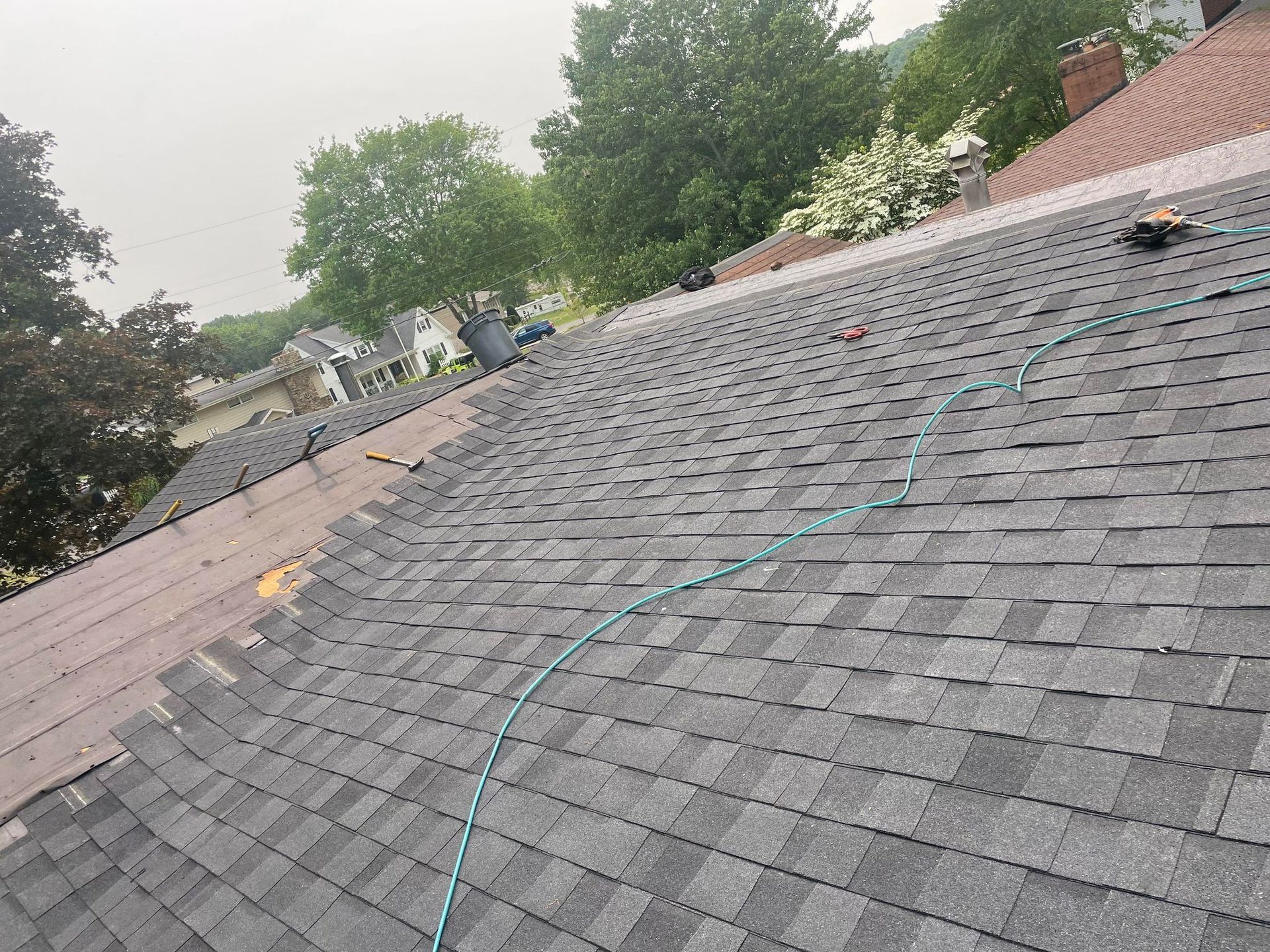 Roof with gray shingles partially replaced, surrounded by trees and houses.