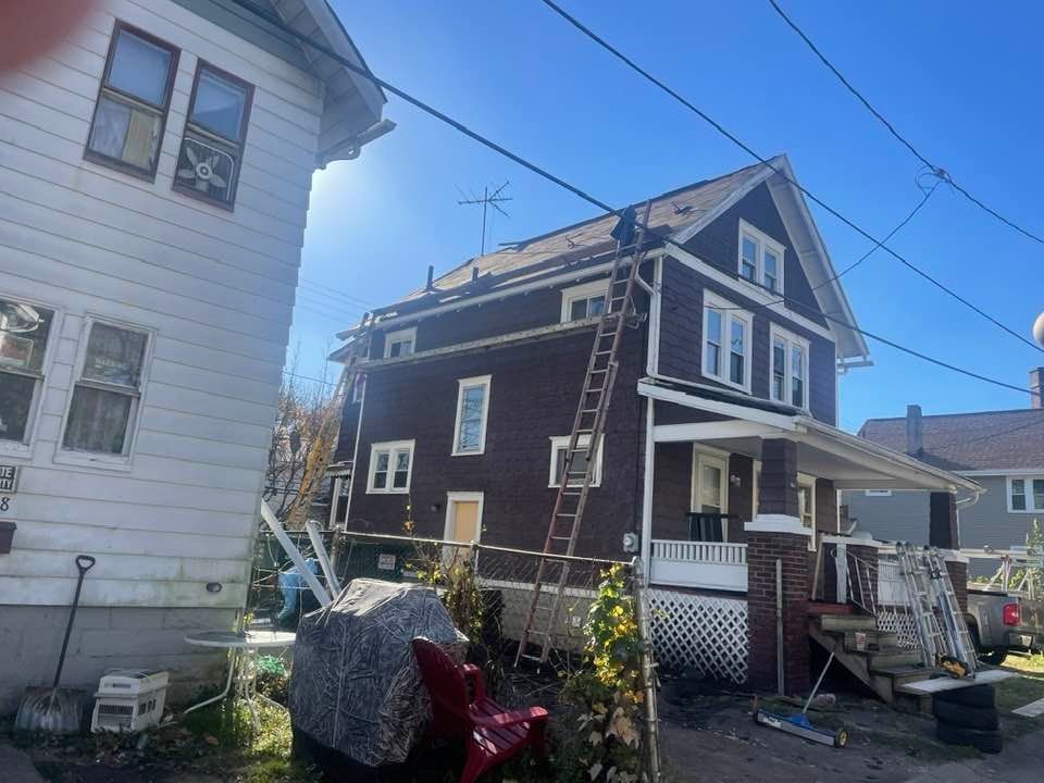 Two-story brown house with a ladder against the roof, next to a white house under a blue sky.