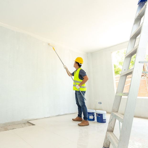 Person in safety vest and hard hat painting a white wall with a roller. Ladder and paint buckets visible. Person in safety vest and hard hat painting a white wall with a roller. Ladder and paint buckets visible.