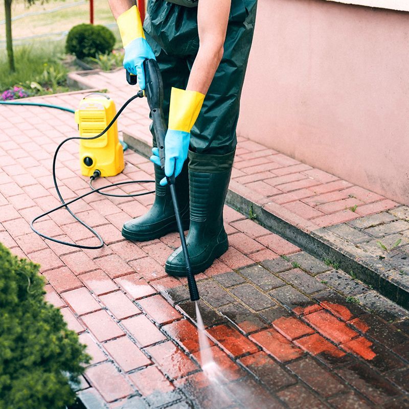 Person power washing red brick pavers. They wear green boots and rain pants, blue gloves, and yellow gloves. Person power washing red brick pavers. They wear green boots and rain pants, blue gloves, and yellow gloves.