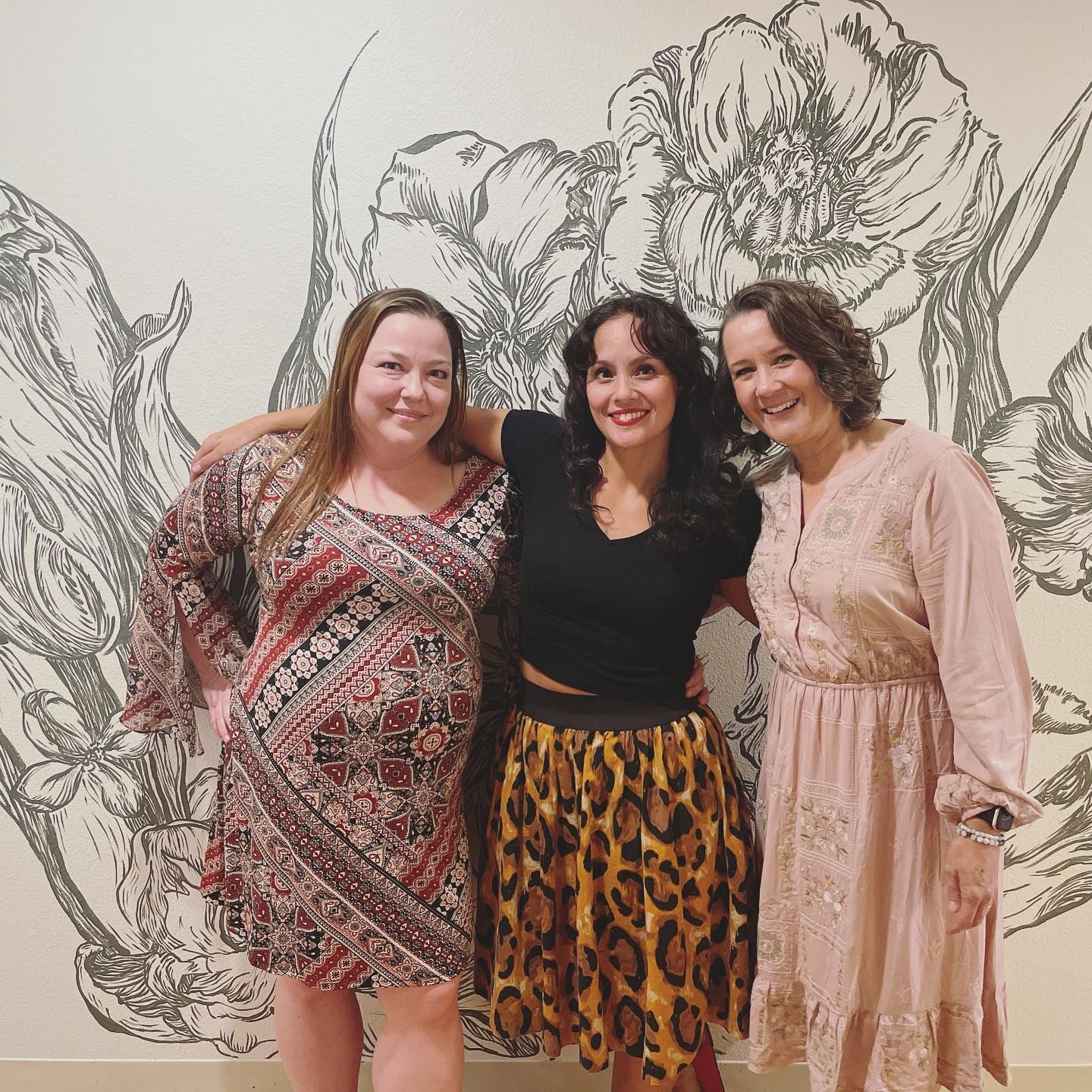 Three women smiling, posing in front of floral wall art. One wears a leopard-print skirt.
