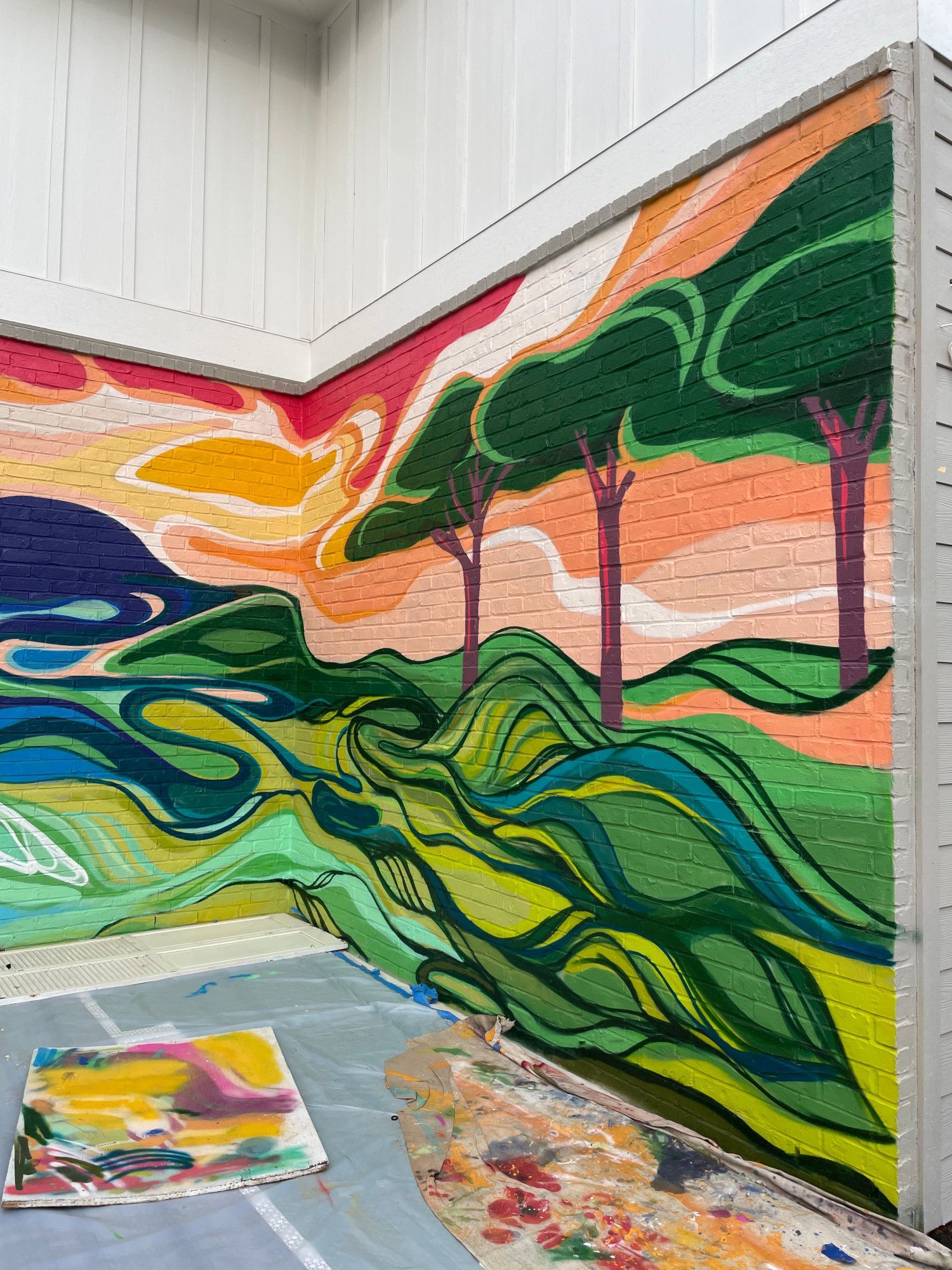 Mural on a brick wall depicts a colorful landscape with trees, rolling hills, and a swirling river; an artist's palette and canvas lay on the ground.