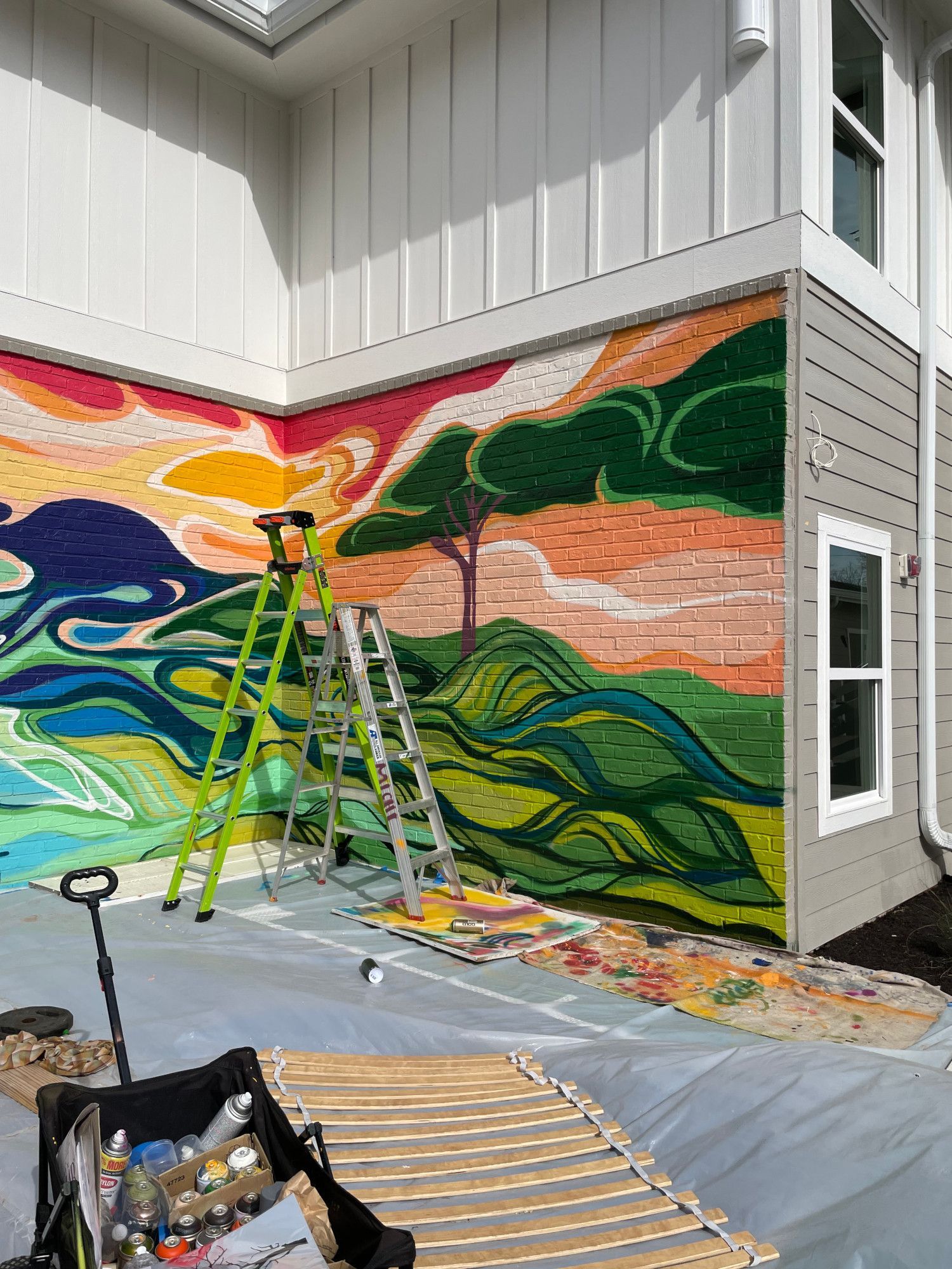 Mural in progress: colorful landscape on a building exterior. Ladders, paint supplies, and artist's chair present.