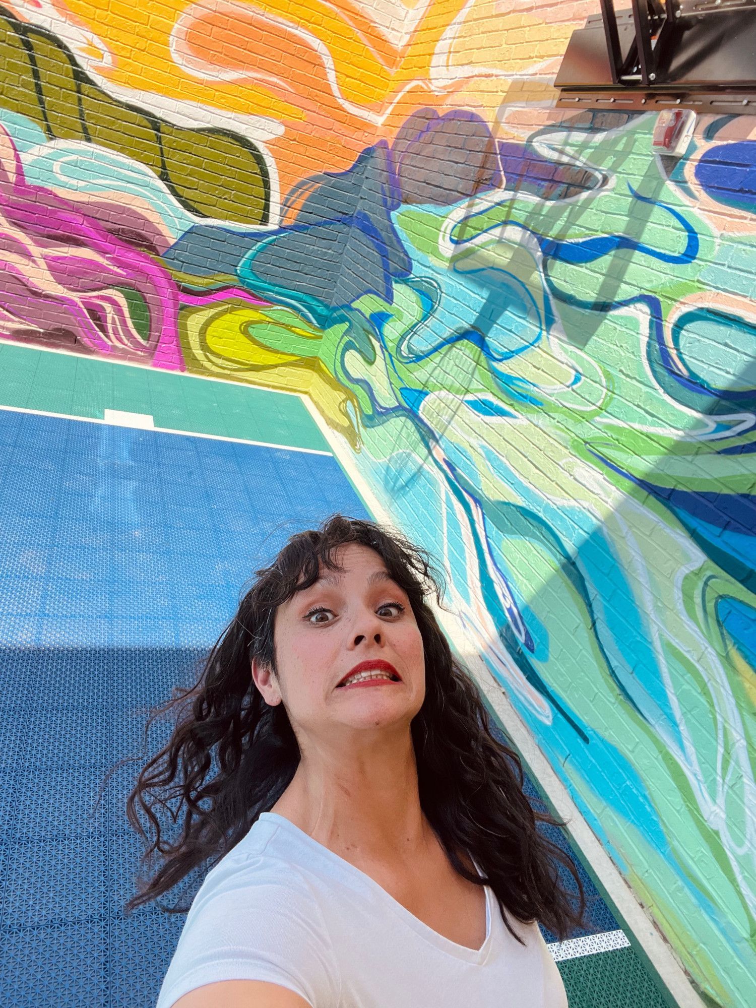 Woman poses with a colorful mural. She has dark hair, wears a white shirt, and makes a surprised face.