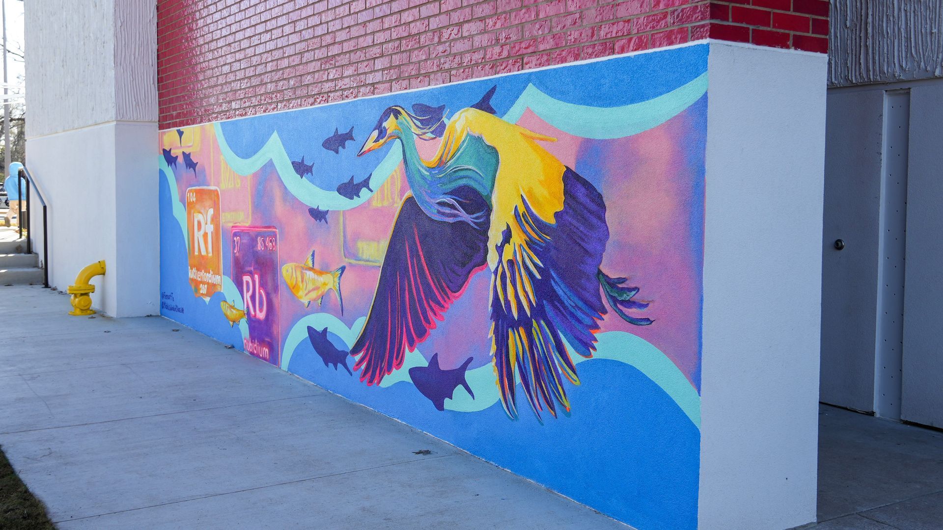 Mural with a large bird and ocean-themed elements, painted on a textured blue wall next to a sidewalk.