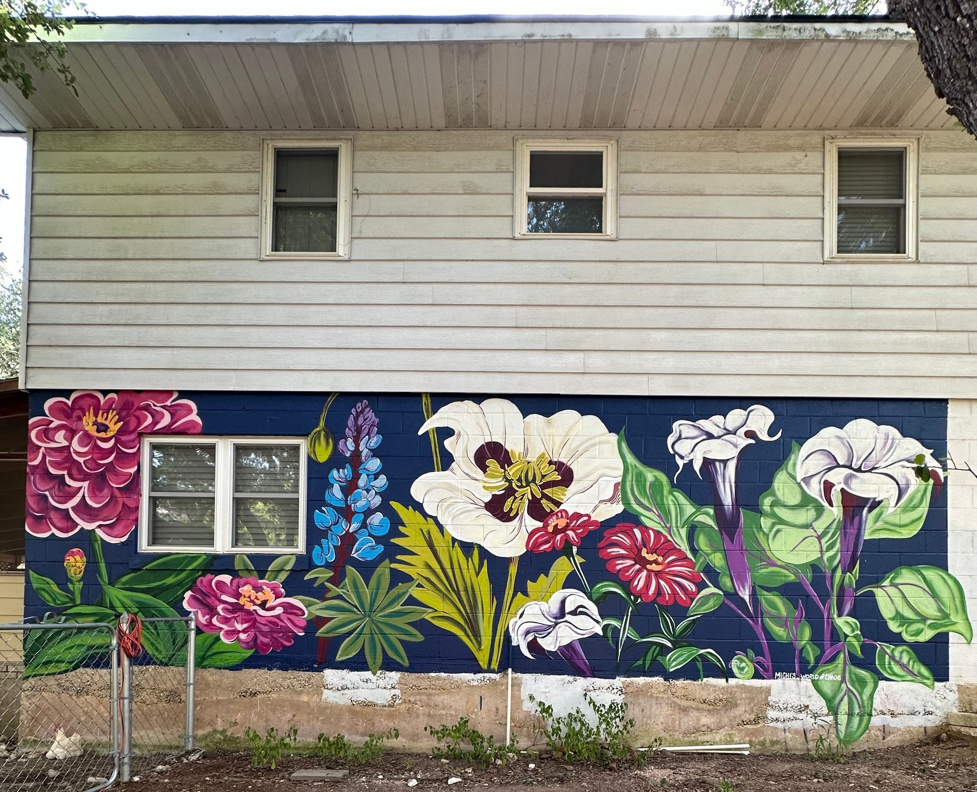 Mural of vibrant flowers on a blue background on a white-sided building. Three windows above.