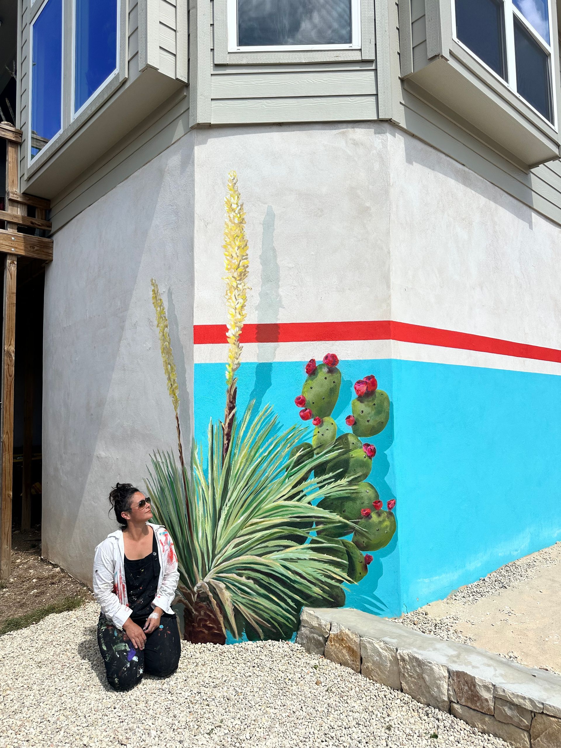 Woman kneeling beside a painted mural on a building corner with cactus and desert plants.