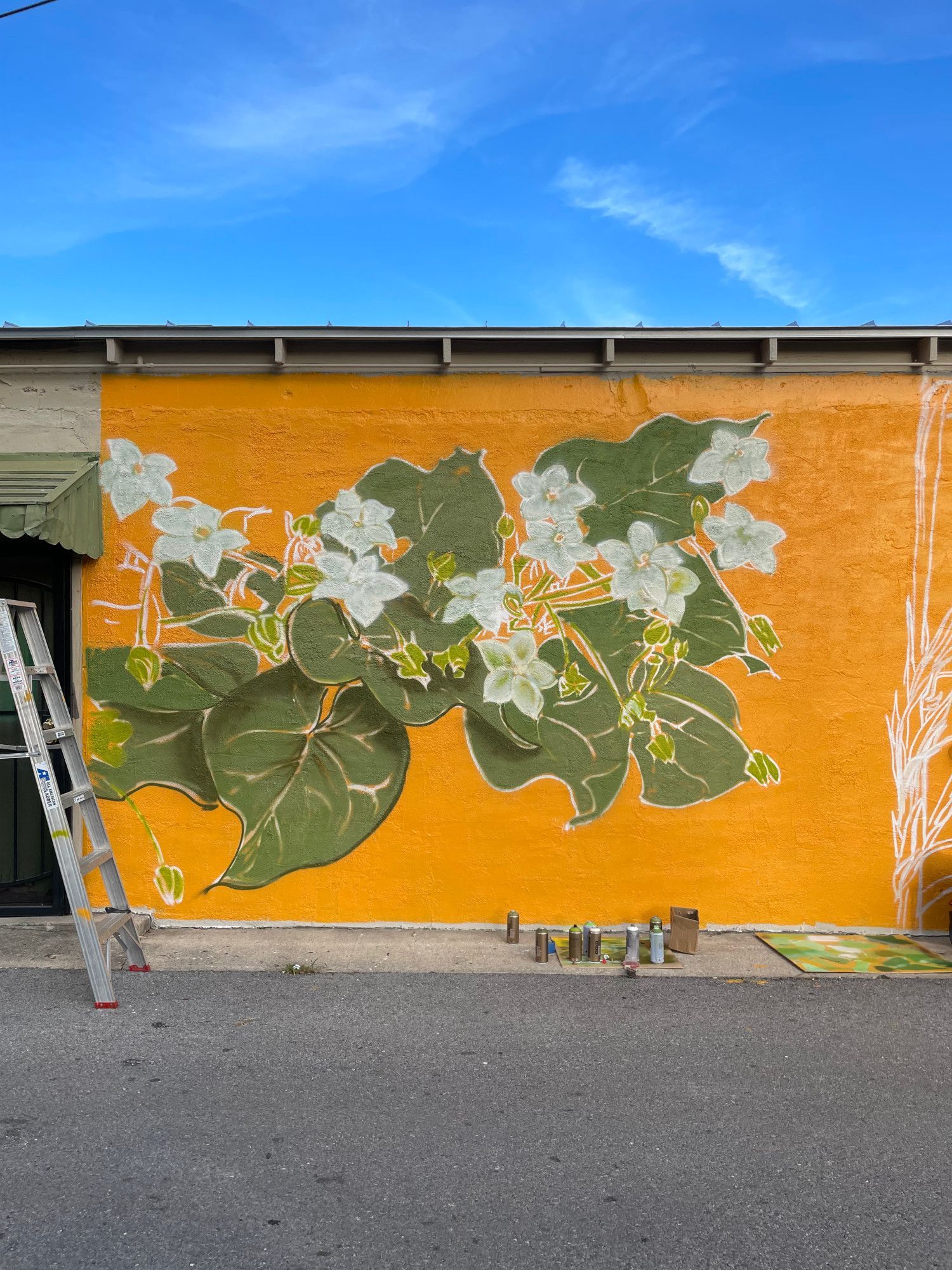 Mural of white flowers and green leaves on a yellow wall. A ladder and spray paint cans are visible.