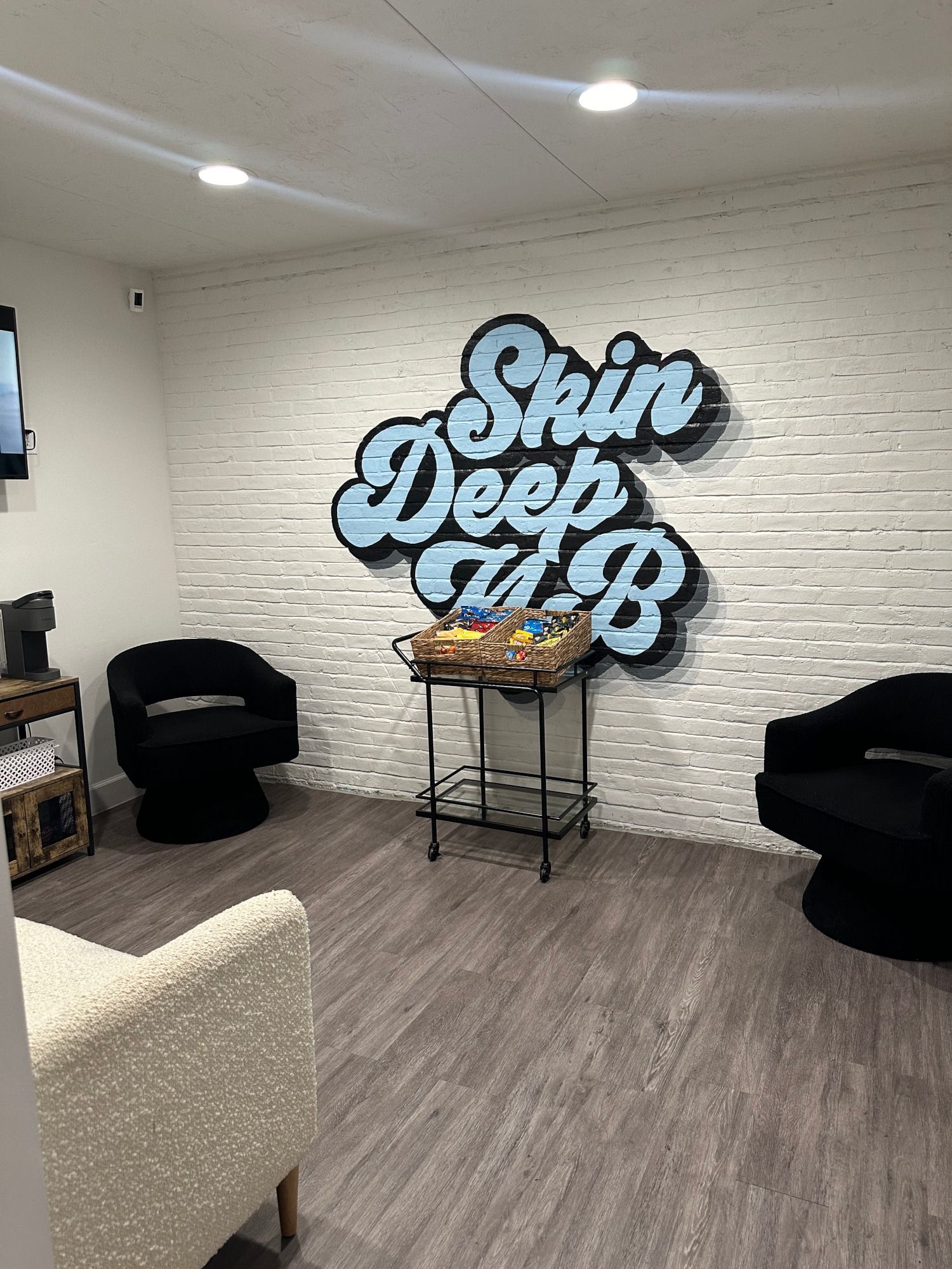 Interior waiting area with Skin Deep A&B logo on a white brick wall; black chairs.