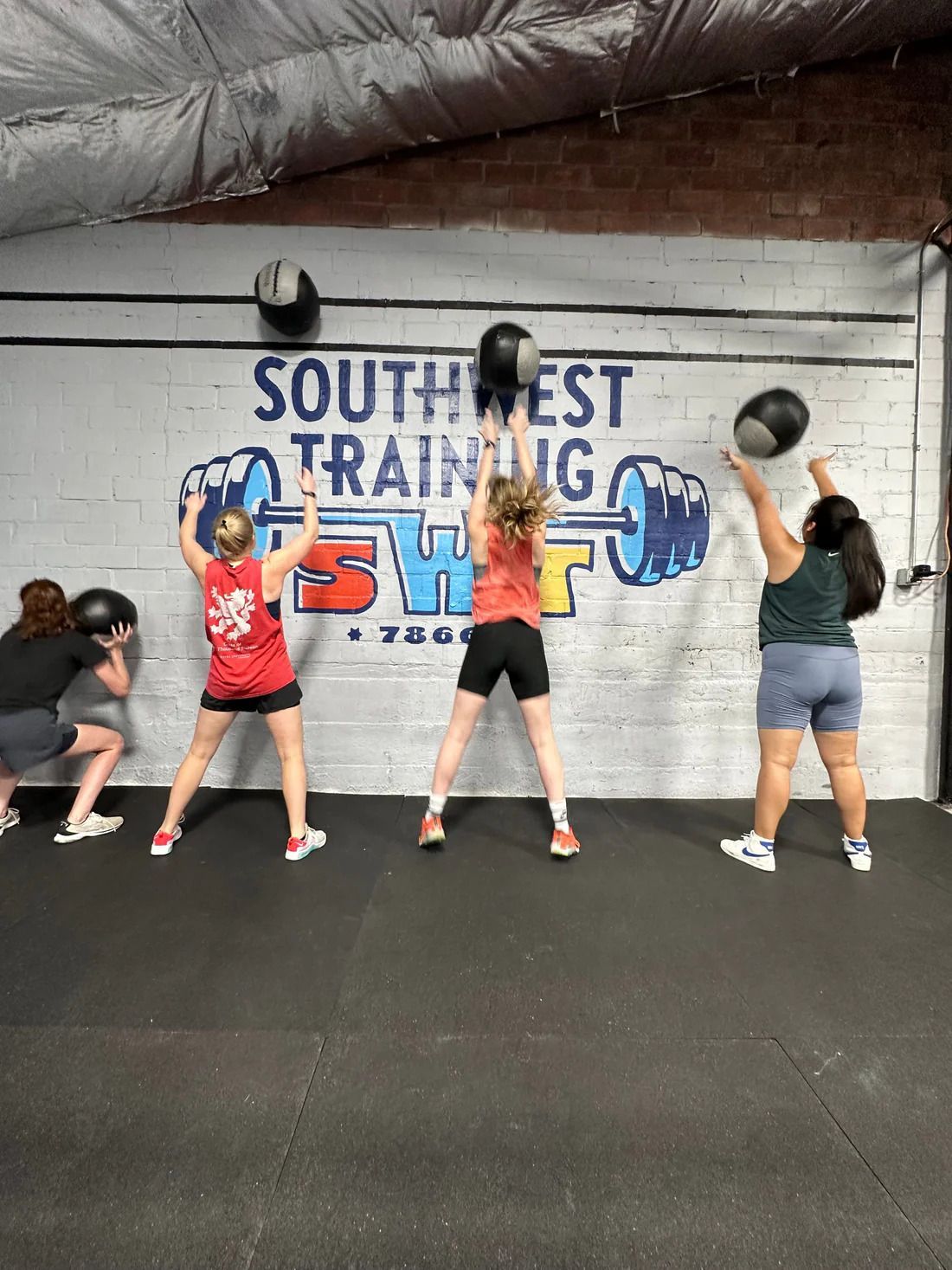 Four women in gym throw medicine balls against a wall that reads