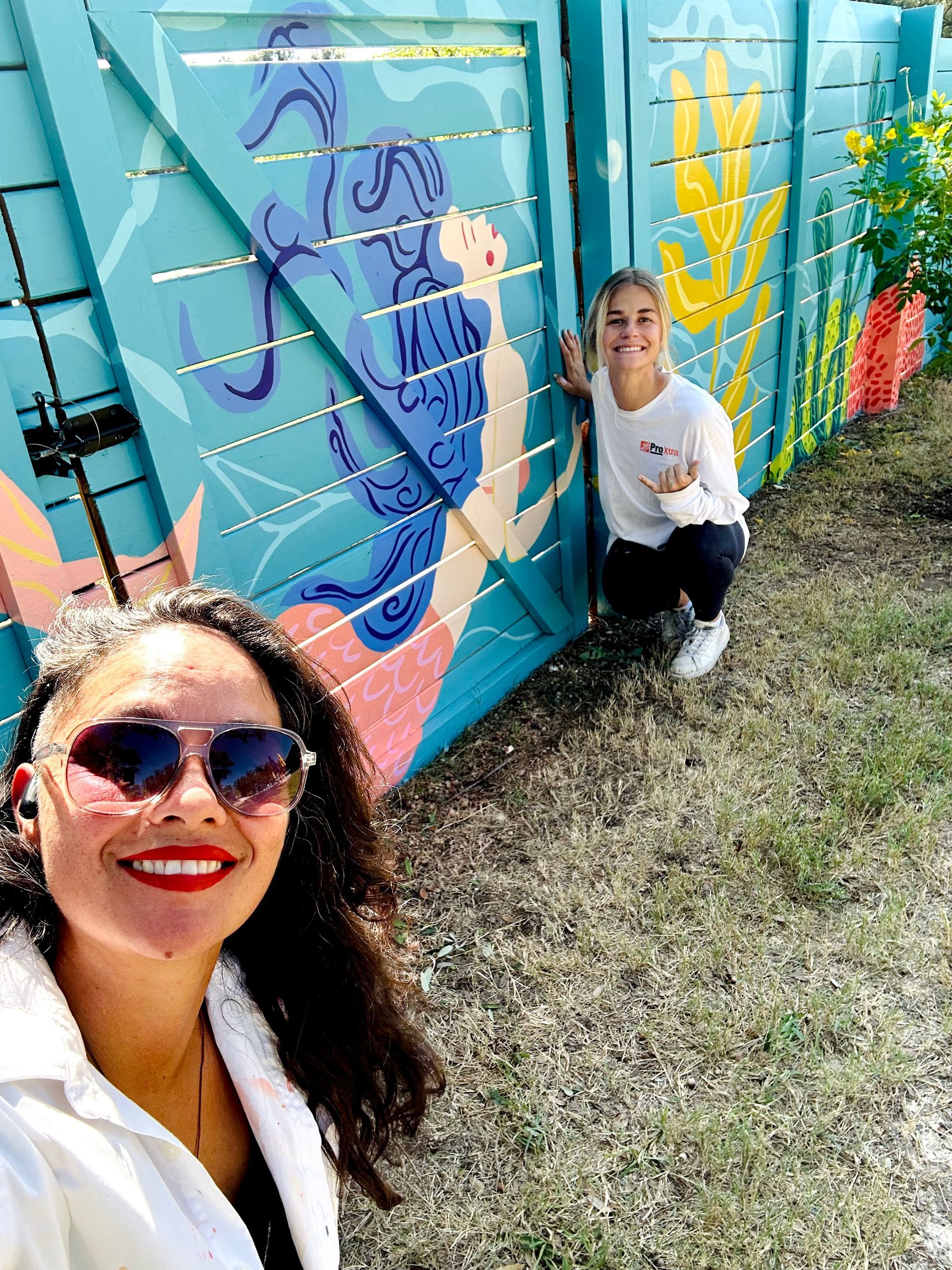 Two people pose by a turquoise fence mural with a mermaid. One takes a selfie, the other smiles.