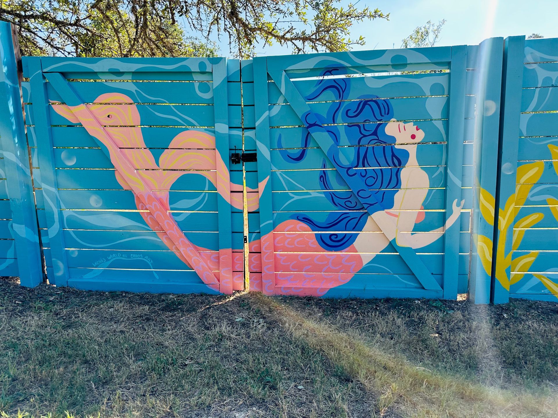 Painted mermaid on a blue wooden fence; blonde skin, pink tail, blue hair, underwater scene.