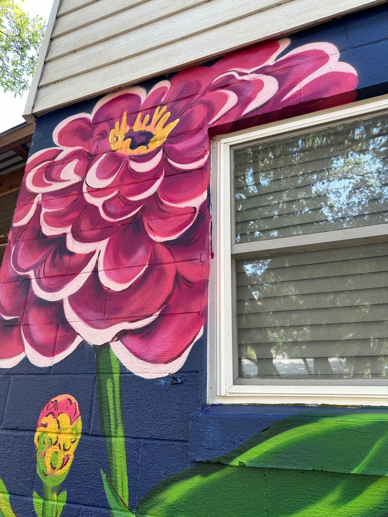 Mural of a large magenta flower on a blue brick wall, next to a window.