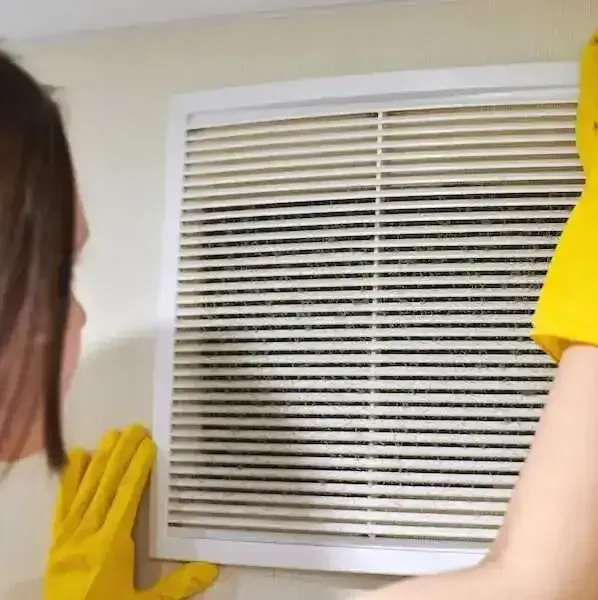 A woman wearing yellow gloves is cleaning a vent on a wall.