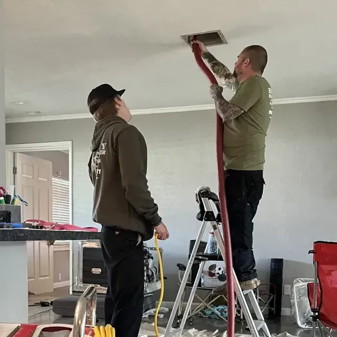 Two men are working on a ceiling vent in a living room.