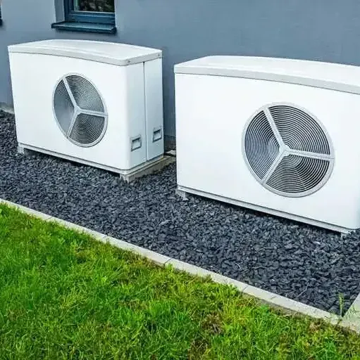Two air conditioners are sitting next to each other on gravel in front of a building.