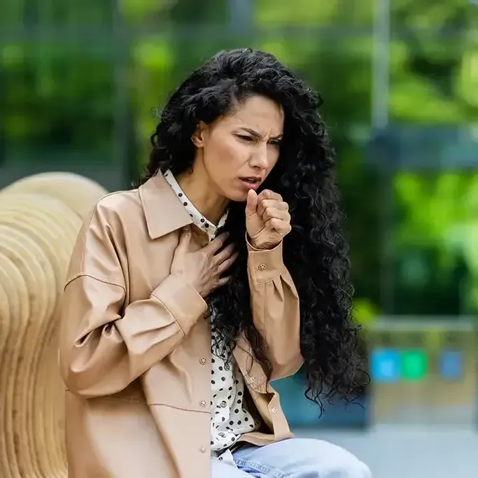A woman with long hair is sitting on a bench and coughing.