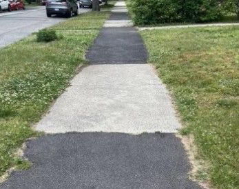 Cracked sidewalk path beside a grassy roadside with parked cars in the distance