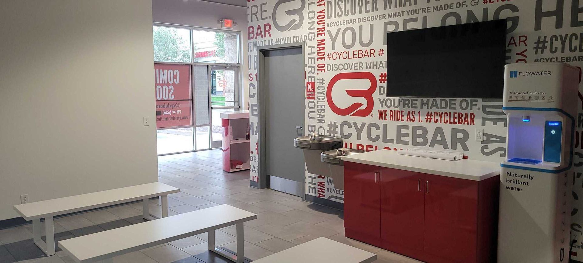 Waiting area with benches, a water cooler, a television, and branded red and white walls at a CycleBar fitness studio.