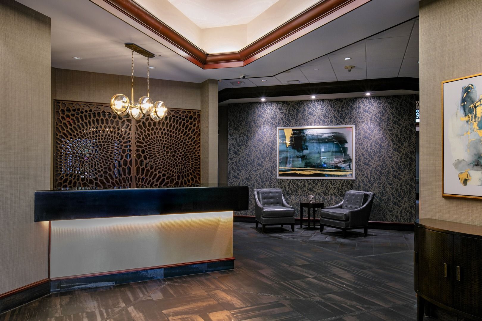 A hotel lobby reception desk with a modern chandelier, decorative screen, patterned wallpaper, and two gray armchairs.