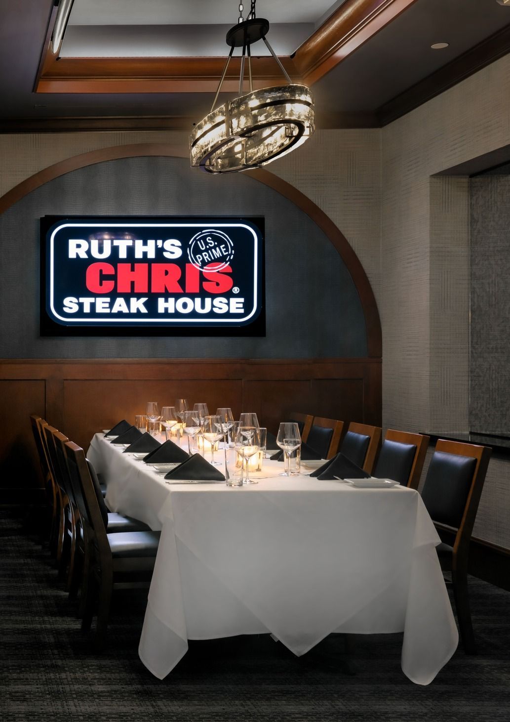 A white-clothed long dining table set for a formal dinner under a chandelier, with a Ruth's Chris Steak House sign behind.
