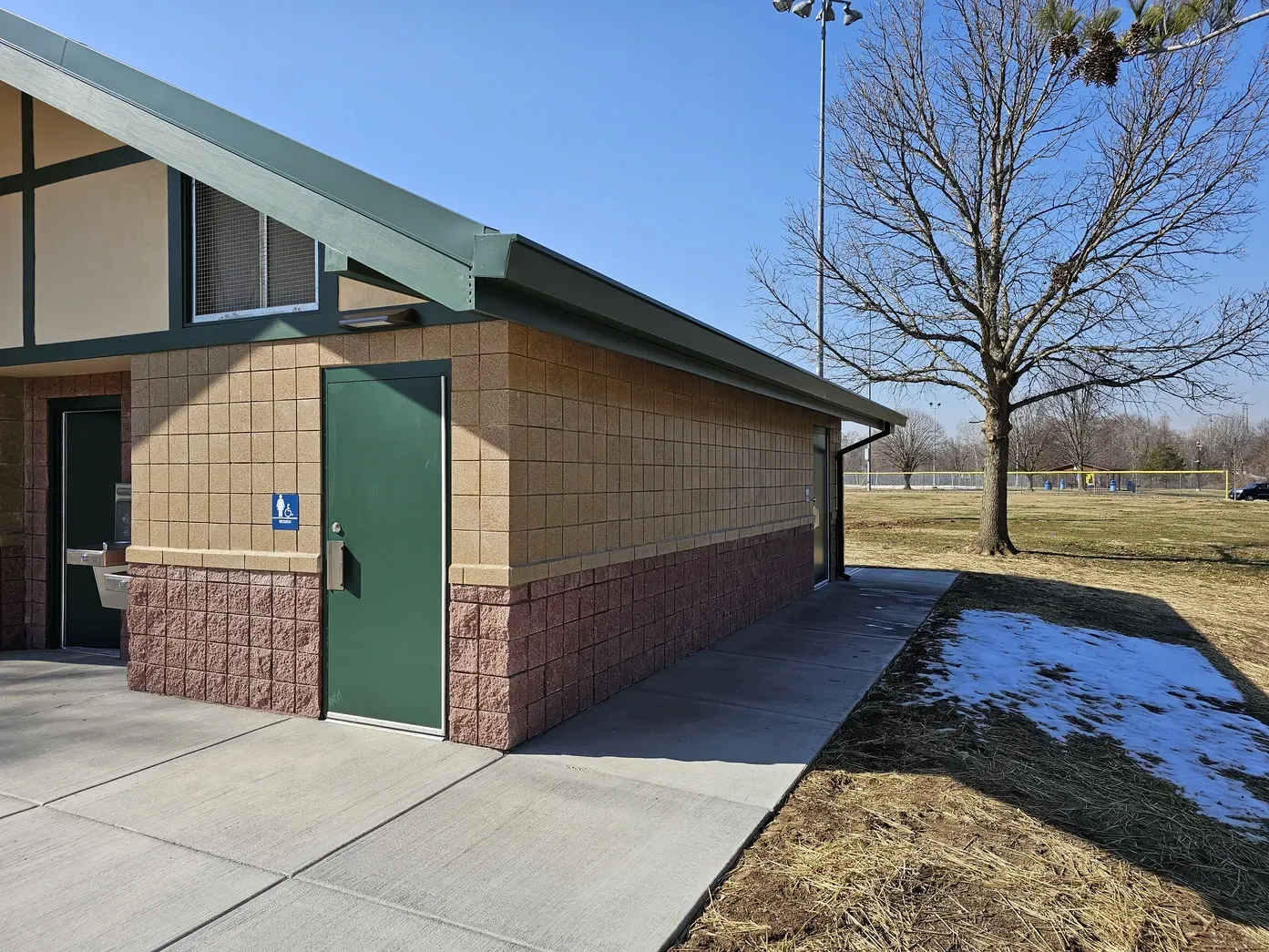 A tan brick building with green doors and roof at a park with a concrete walkway and patches of snow on the ground.