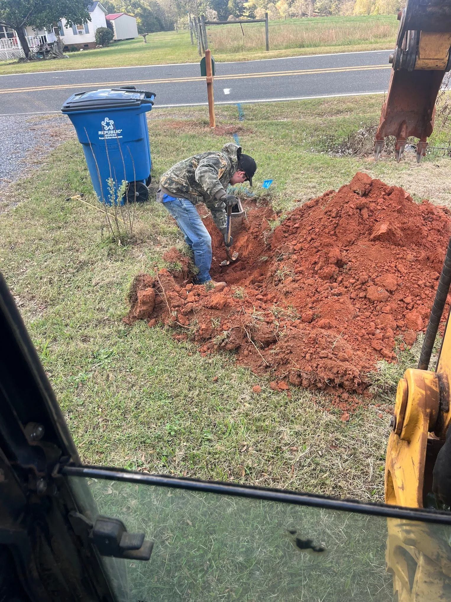 Person digging a mound of red dirt beside a blue trash bin near a roadside, viewed from inside a vehicle