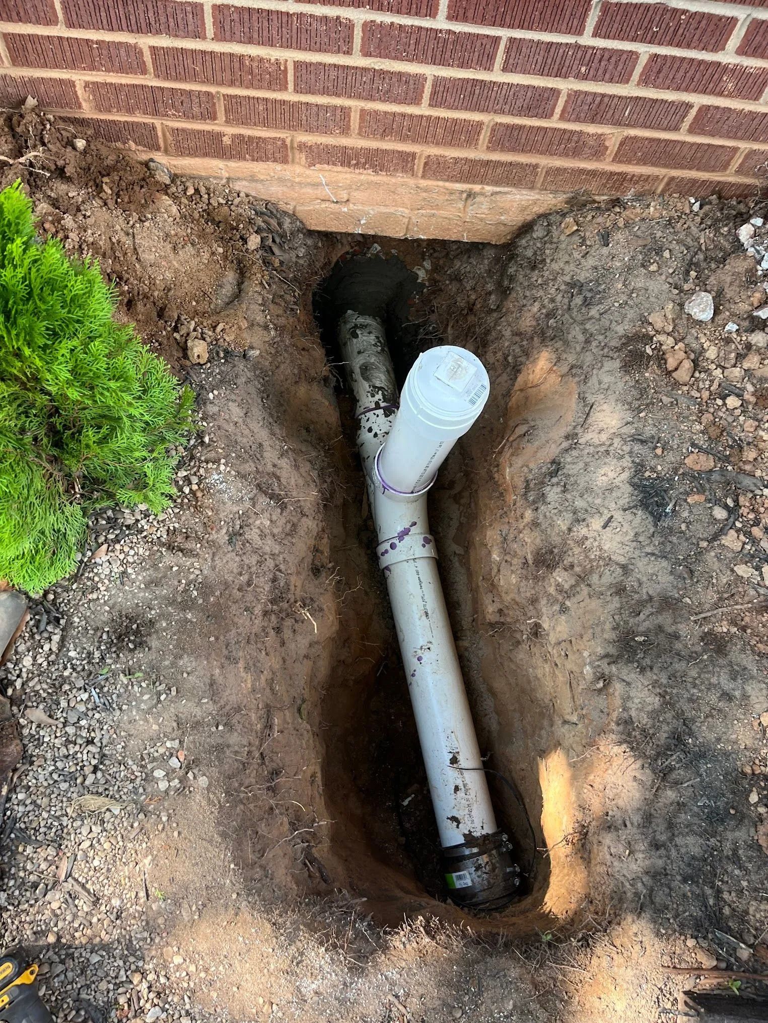 Exposed white PVC pipe in a dirt trench beside a brick wall, likely a plumbing repair.