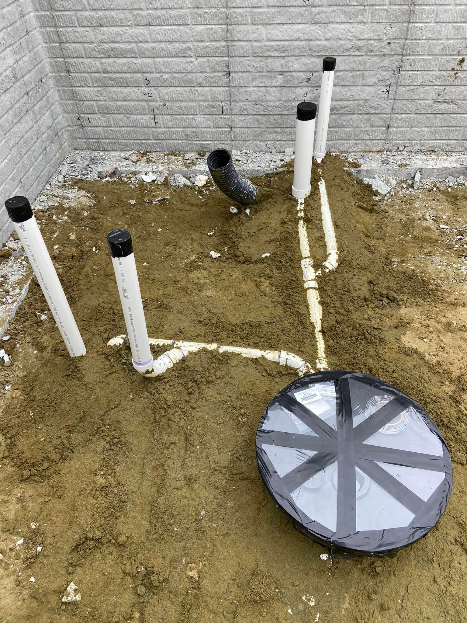 Outdoor utility setup with white PVC pipes, a round lid, and a small bird on dirt beside a concrete wall