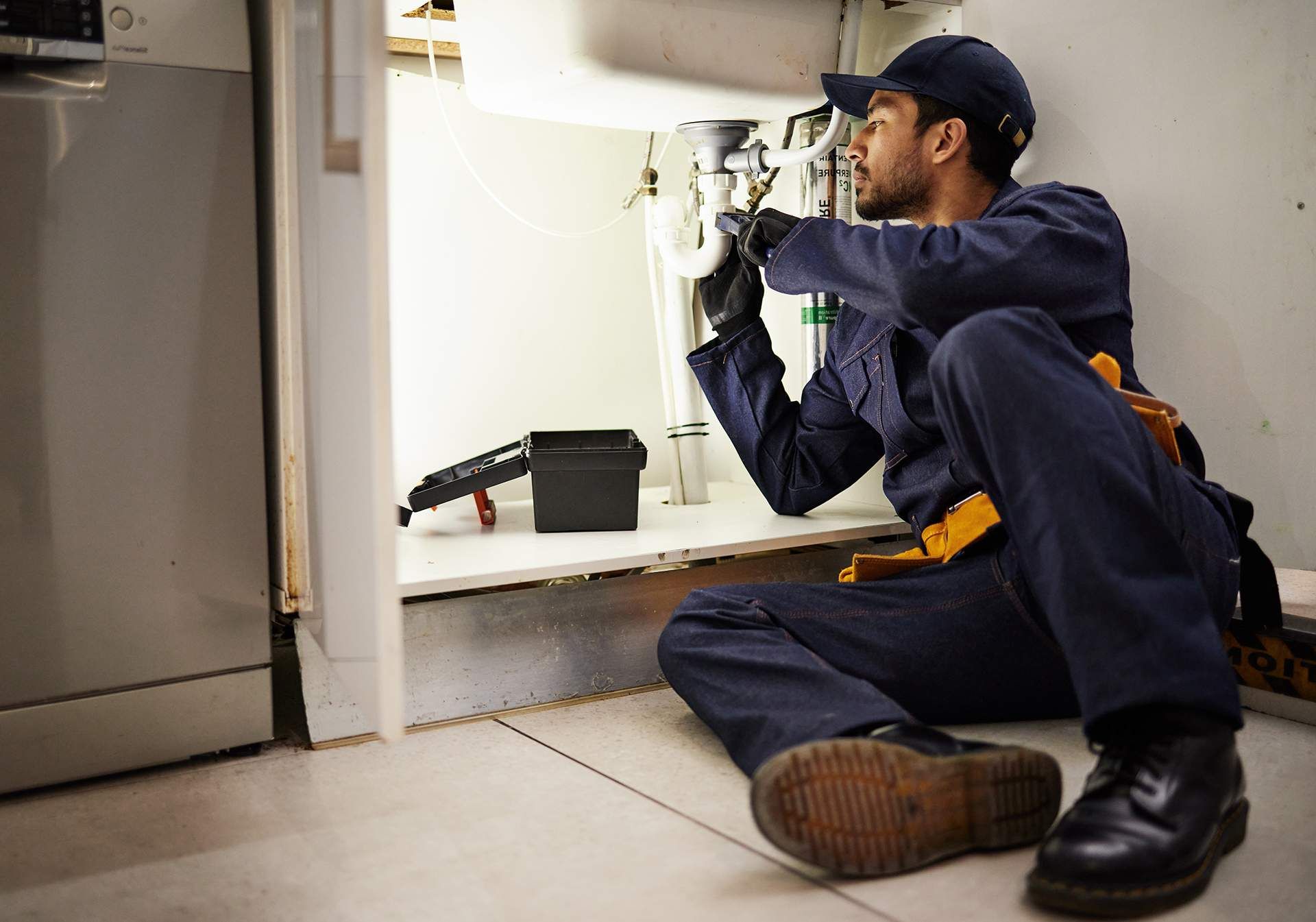 Plumber in blue coveralls inspecting pipes under a sink with a flashlight and tool bag nearby