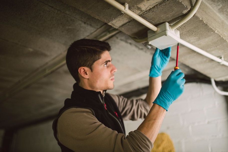 Person in blue gloves adjusts a ceiling light fixture with a screwdriver in a basement.