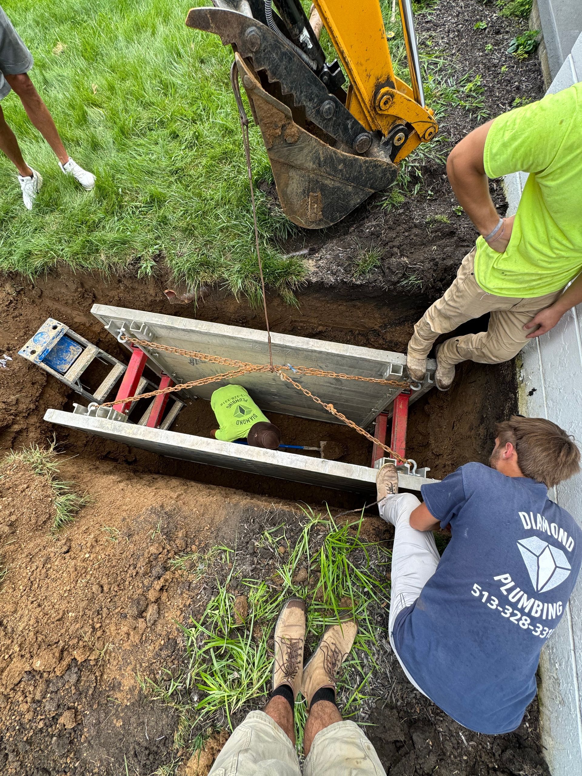 Workers digging a long trench beside a yellow excavator, with tools laid along the opening.