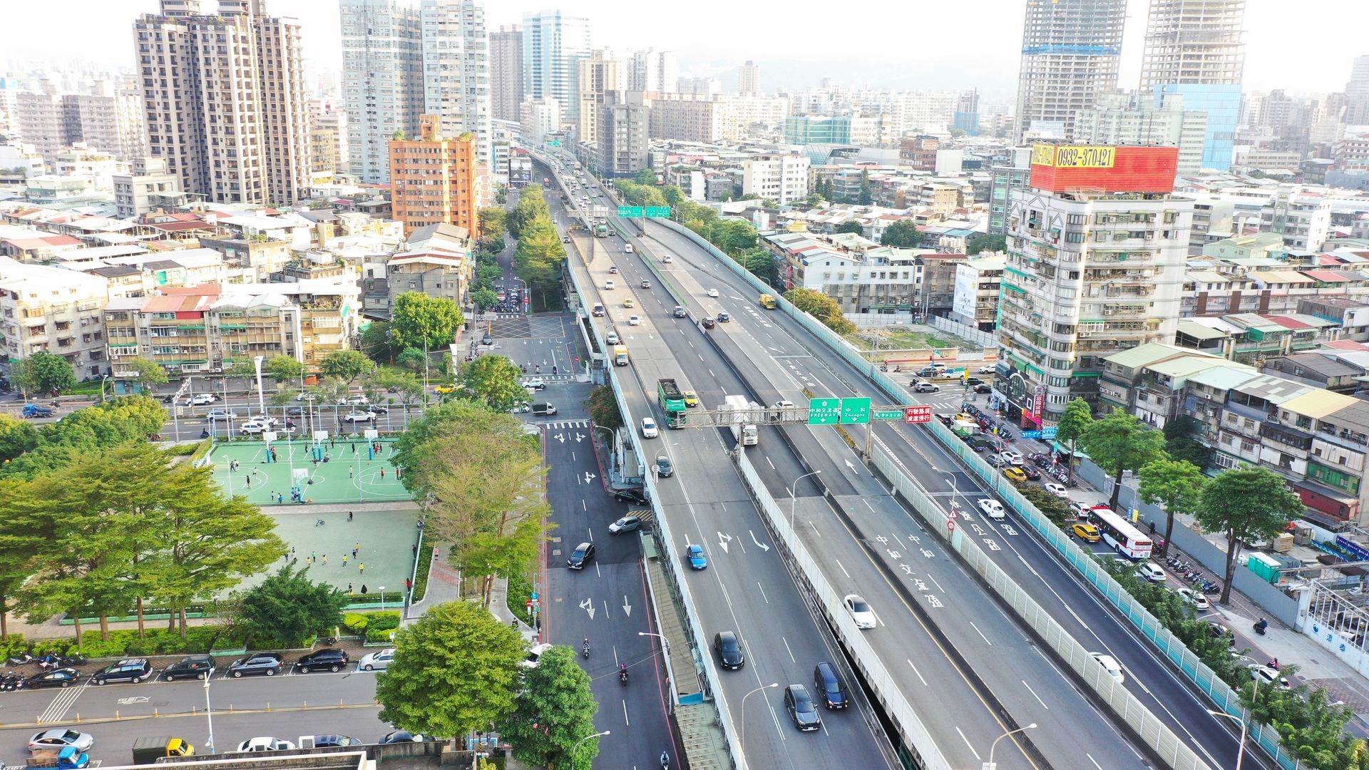 從空中俯瞰繁忙的城市高速公路，兩側是汽車、高樓大廈和綠樹成蔭的公園。