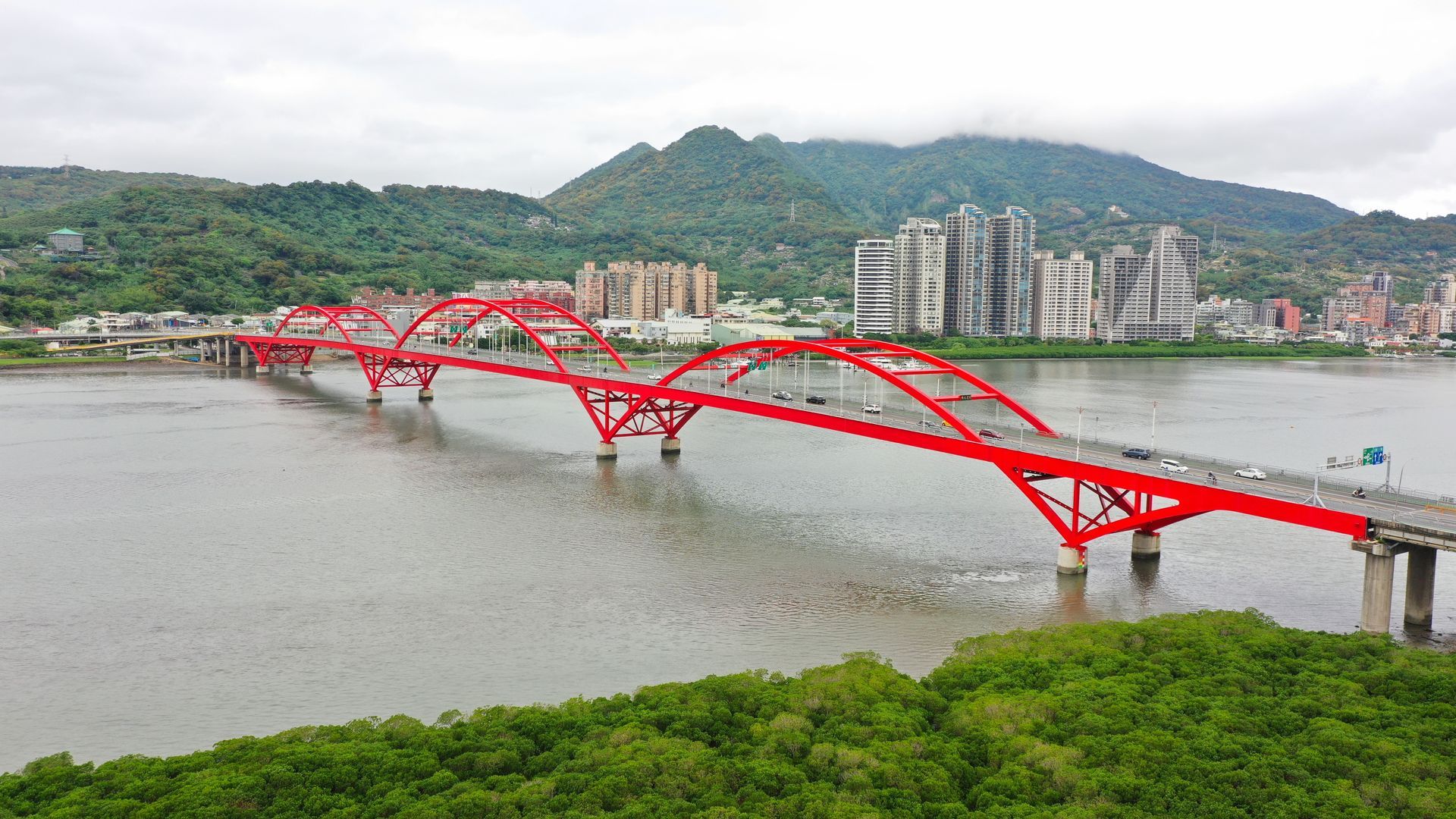 一座紅色拱橋橫跨河流，背景是城市建築和綠色山丘。