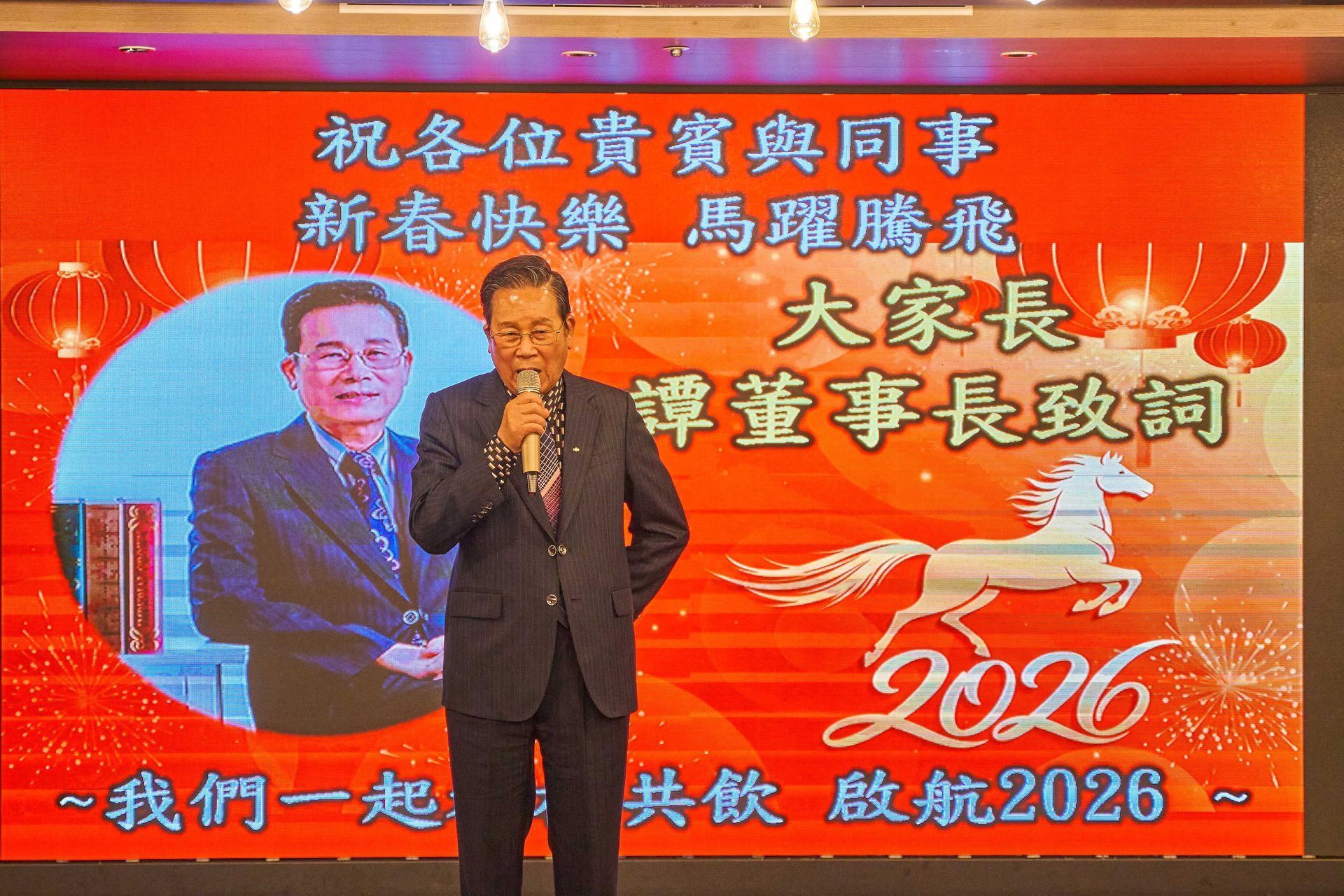 一名男子在台上講話,台前是2026年中國新年紅色背景,背景印有肖像和馬的圖案。