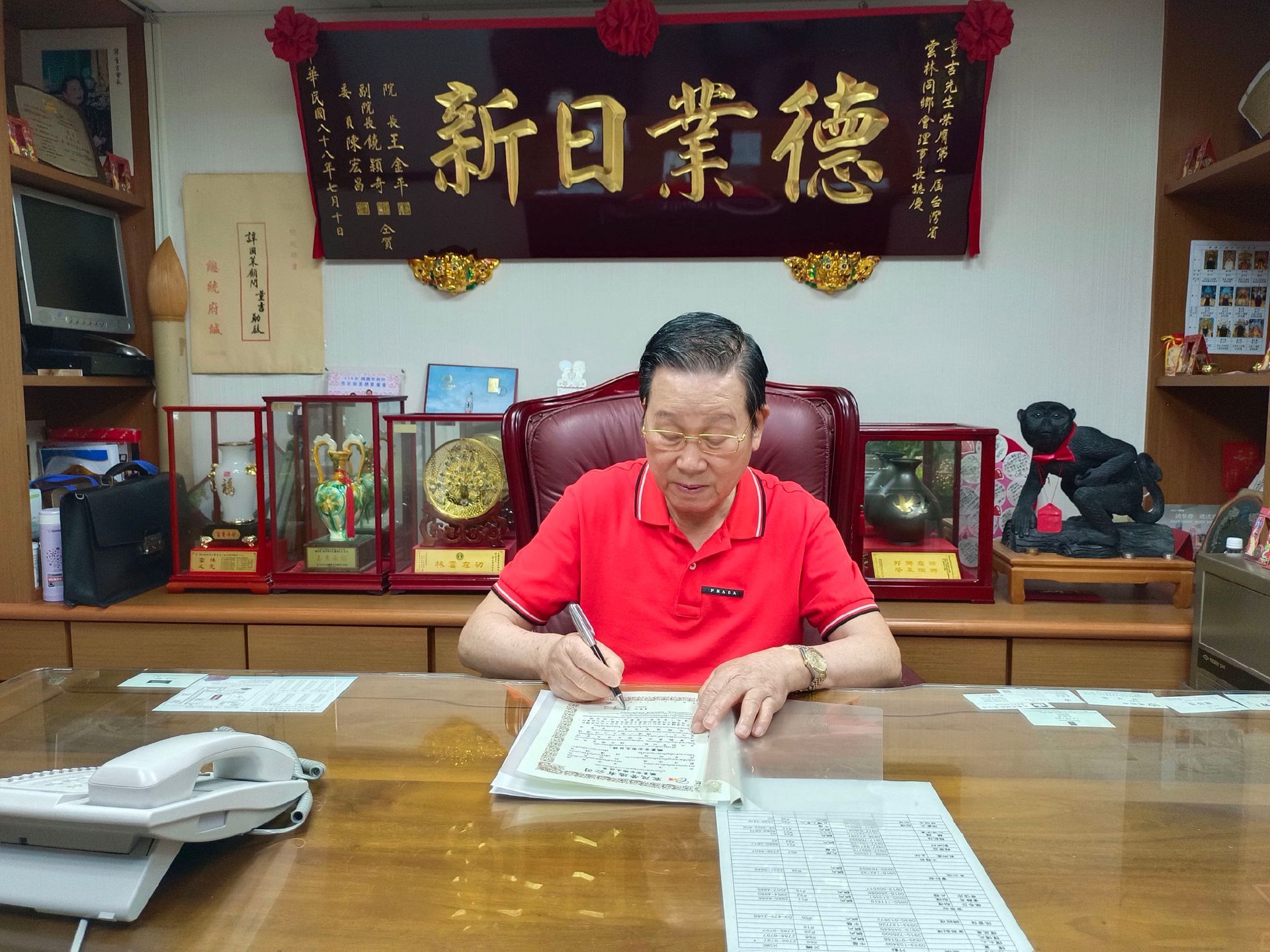 一個穿著紅襯衫的男人正在房間裡的桌子前寫作，房間裡擺放著書法作品牌匾和展示櫃。