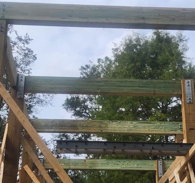 Wooden beams of a structure with green-treated and natural wood, metal brackets, and sky background.
