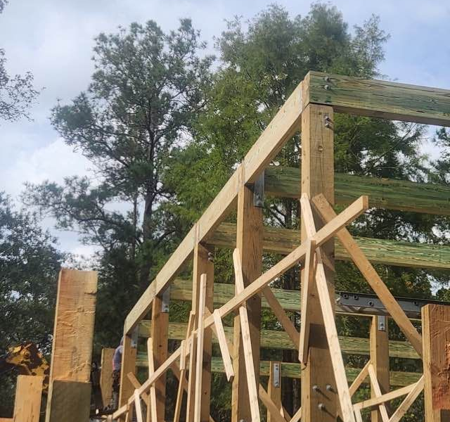 Wooden framework of a building under construction, set against a background of trees and a cloudy sky.