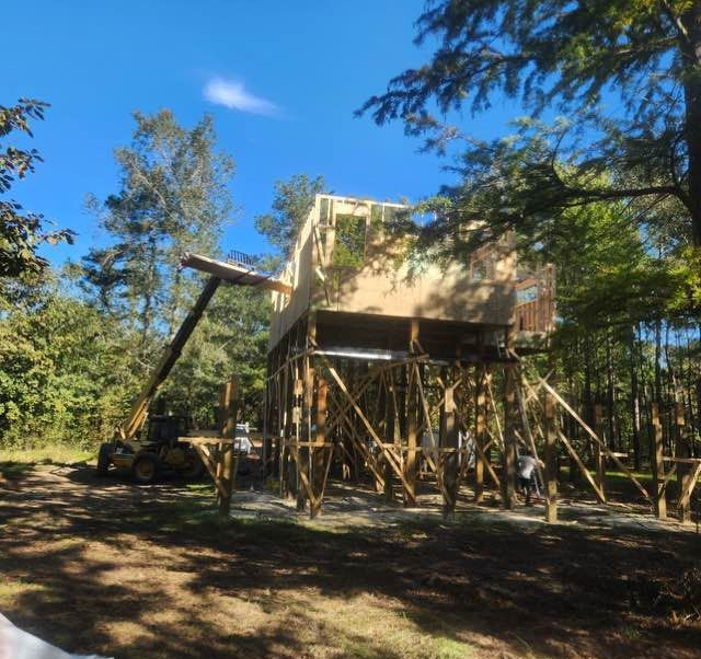 Construction of a raised wooden structure, possibly a treehouse, supported by beams in a wooded area under a blue sky.