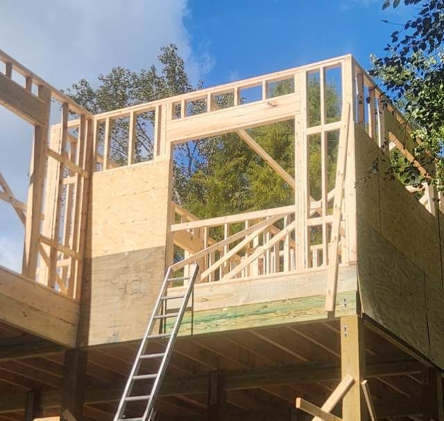 Wooden house under construction with framing and ladder, set against blue sky and trees.