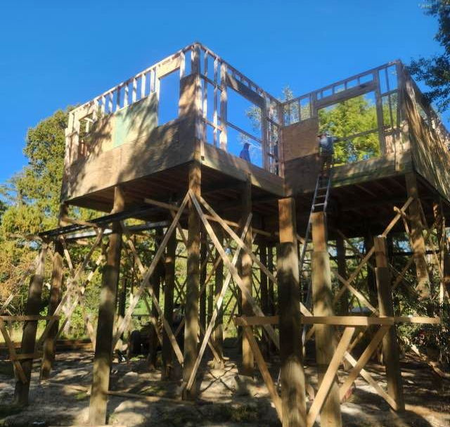Wooden treehouse under construction in a forest; two people visible.
