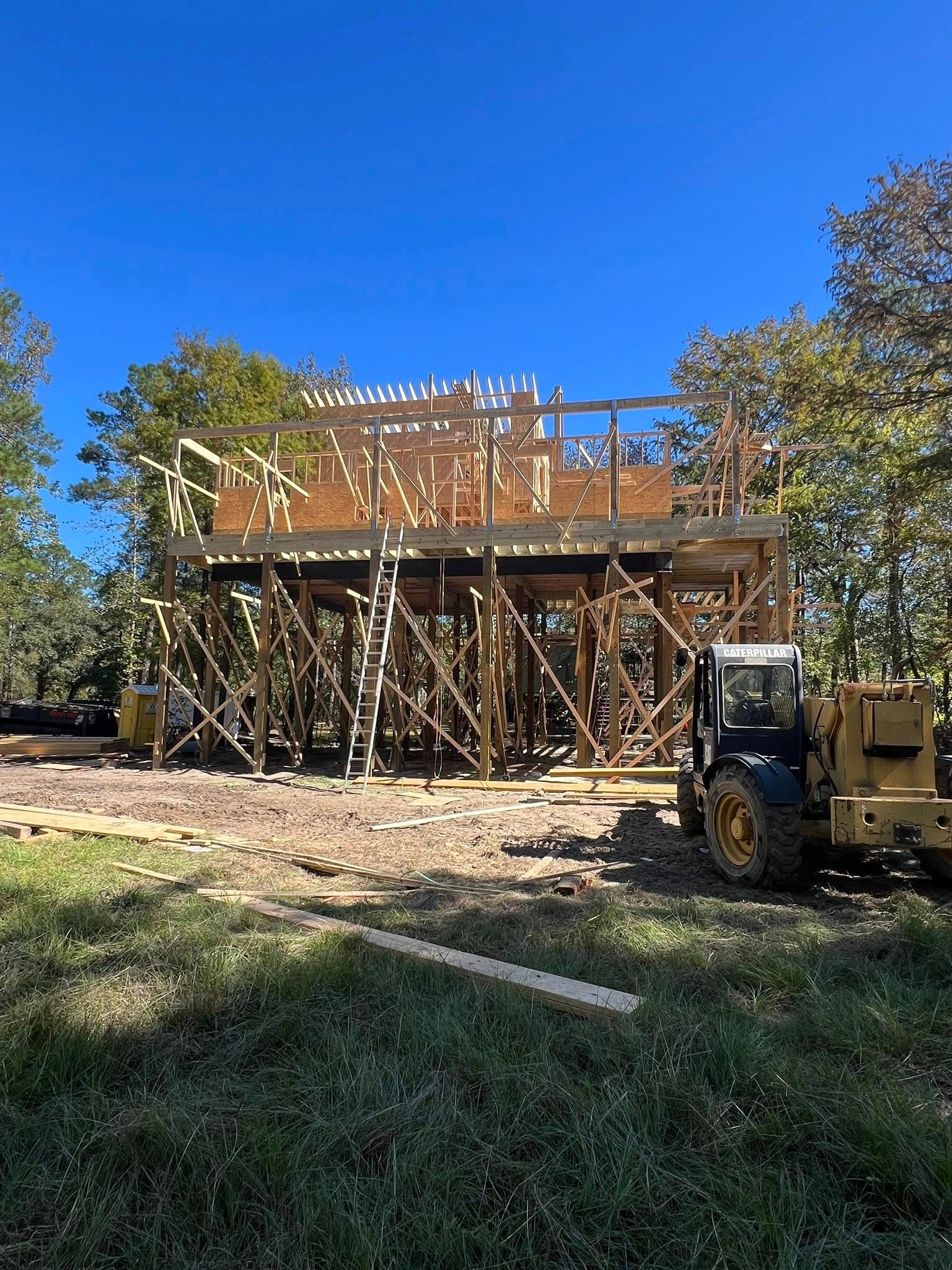Construction site: wooden structure framed, supported by braces, with a yellow loader nearby, under a blue sky.
