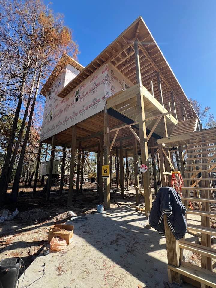 Construction of a house on stilts in a wooded area; wooden frame with wrap, unfinished deck, blue sky.