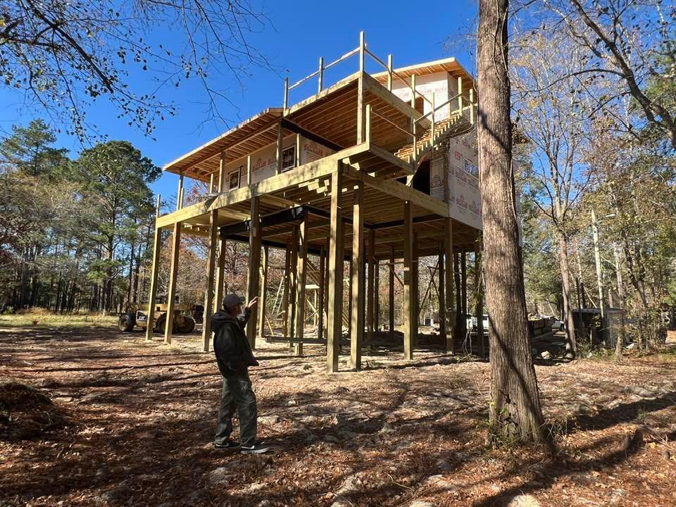 Man stands before a wooden stilt house under construction in a wooded area on a sunny day.