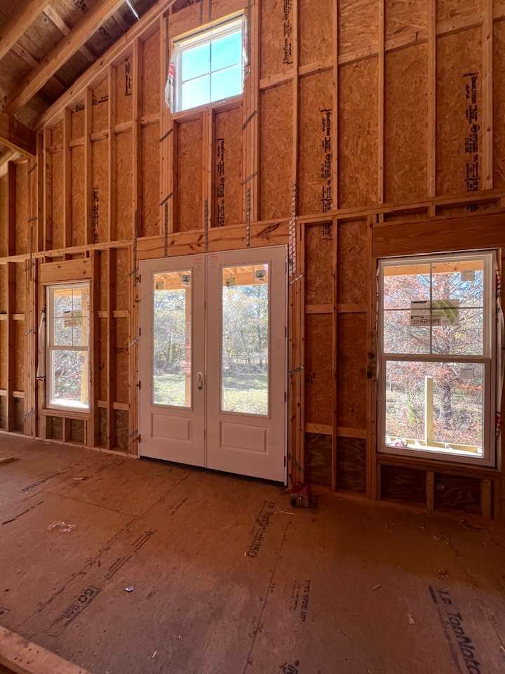 Interior view of a building under construction, showing wooden framing, windows, and a double door.