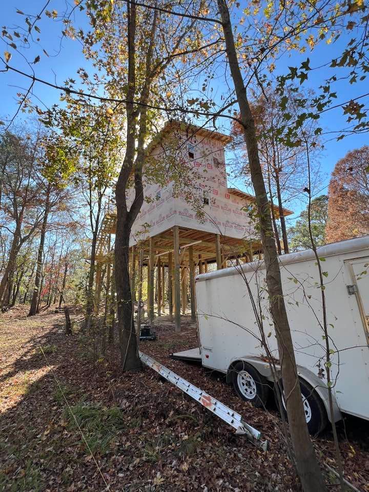 A partially built, elevated wooden structure in a forest, with a trailer parked nearby.