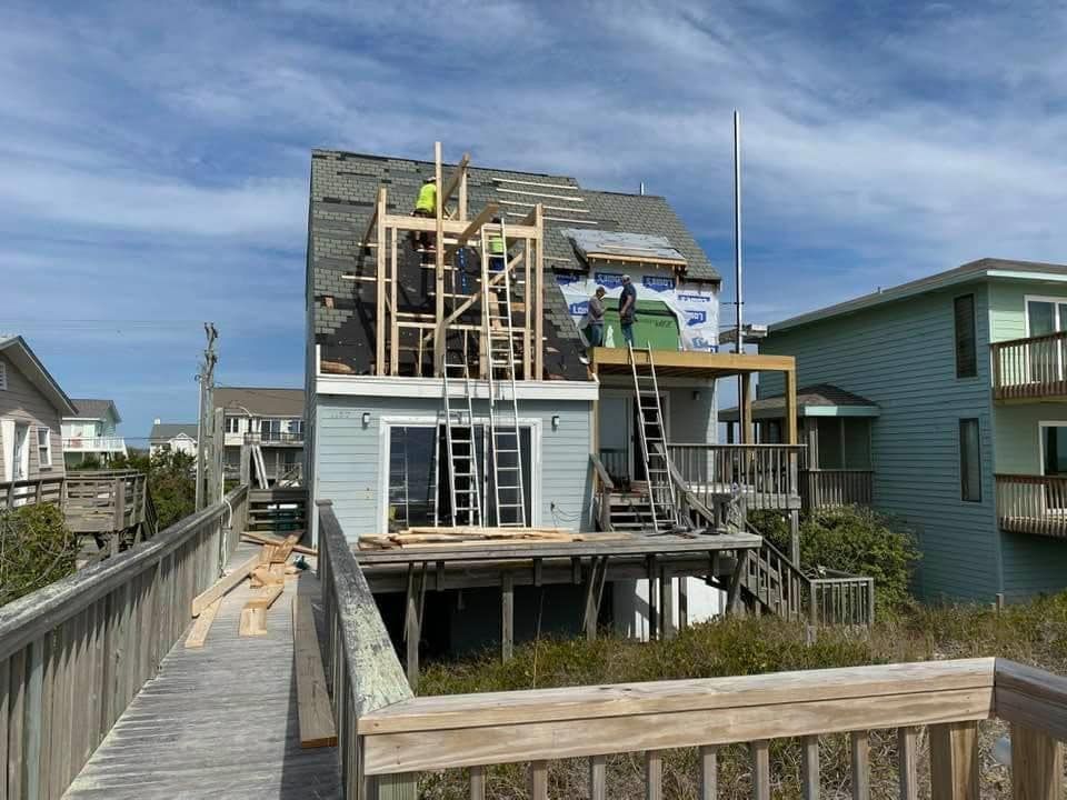 House under construction, roof partially removed; workers on scaffolding; exterior boardwalk, blue sky.